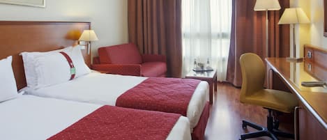 A hotel room with two beds, a desk, a chair, and a small table with a lamp.