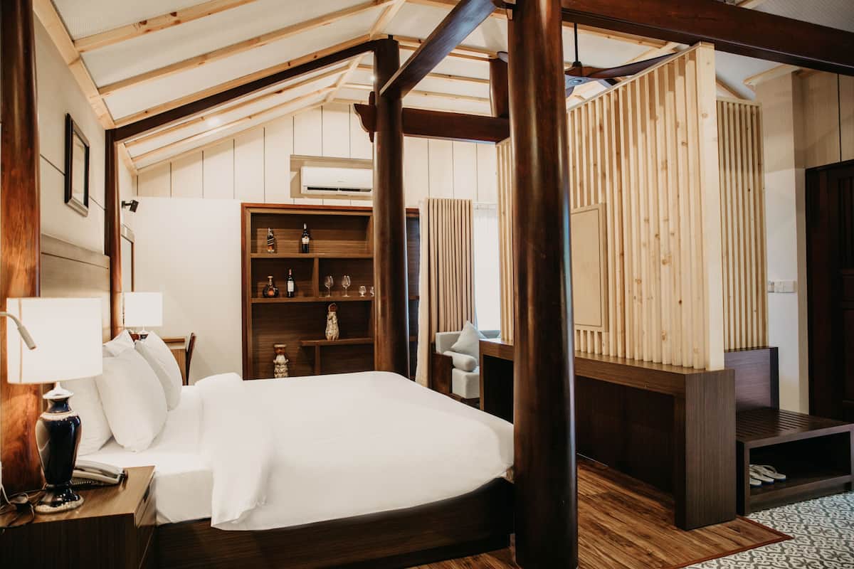 A bedroom with a wooden ceiling, a bed, a nightstand, a lamp, a shelf with bottles, and a chair.