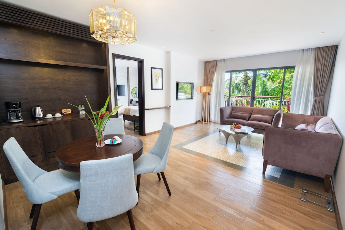 A modern living room with a dining area, a sofa set, and a large window with a view of greenery.