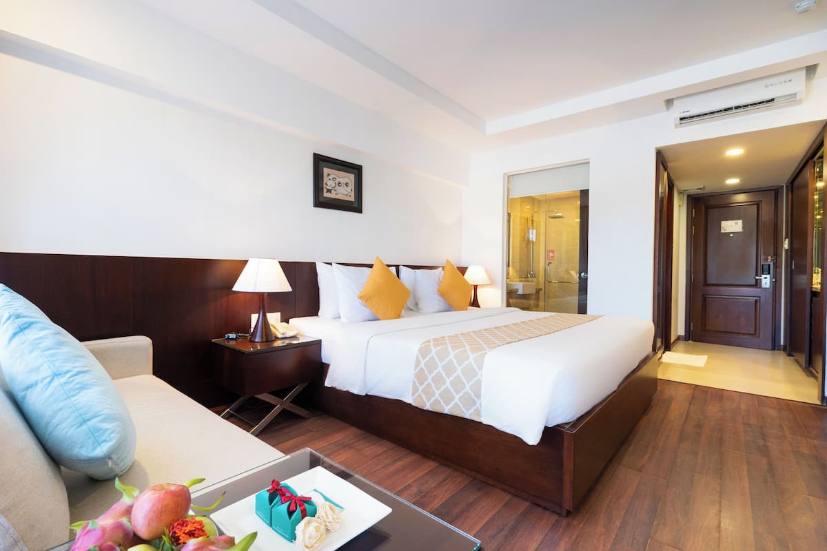 A hotel room with a large bed, bedside tables with lamps, a sofa, and a small table with a plate of fruit and a gift box.