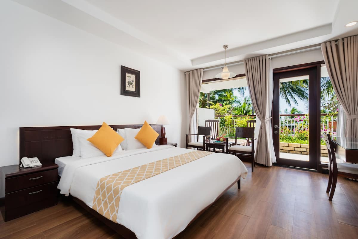 A hotel room with a large bed, a desk with a chair, a phone on the nightstand, and a view of the outdoors through the glass door.