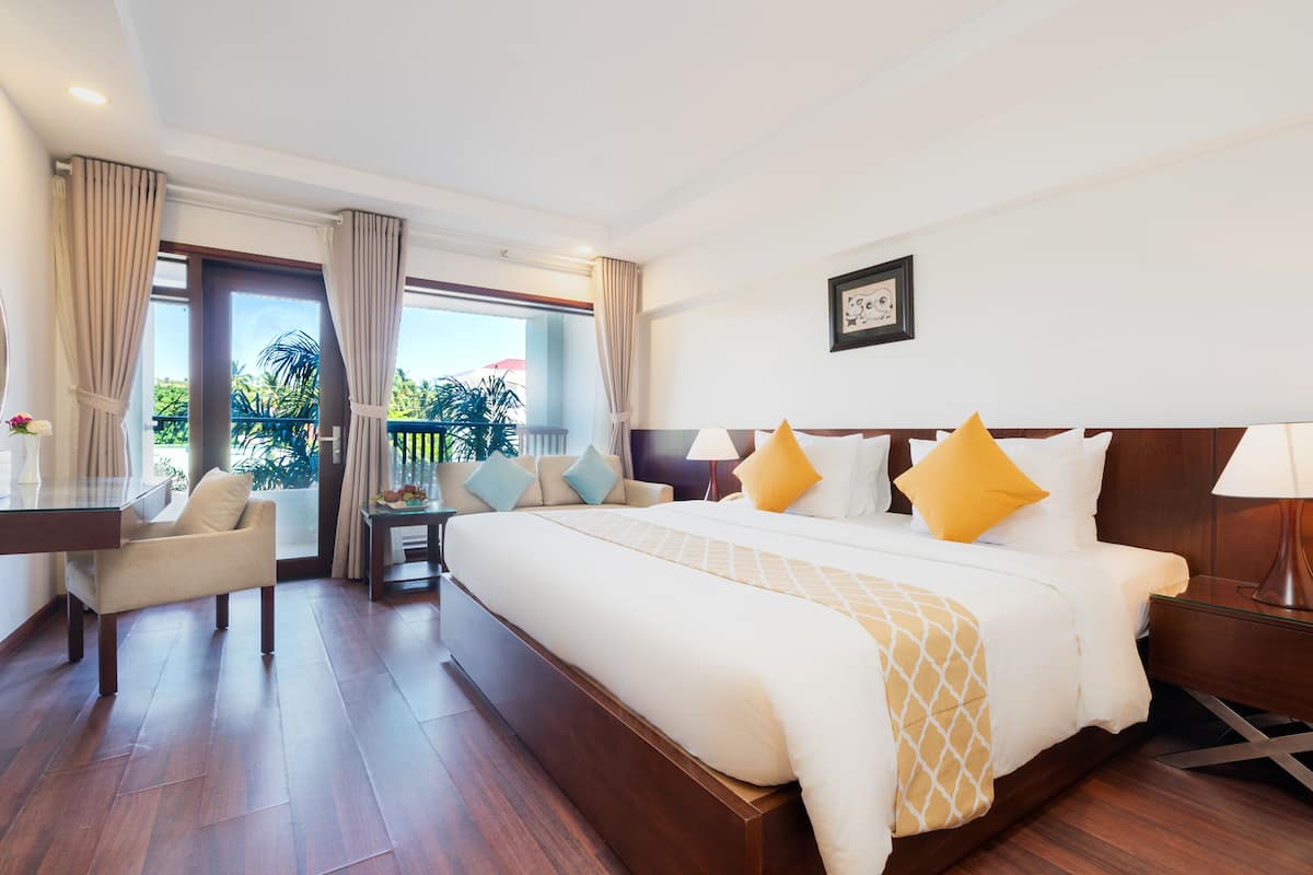 A hotel room with a large bed, a desk, a chair, and a balcony with a view of palm trees.
