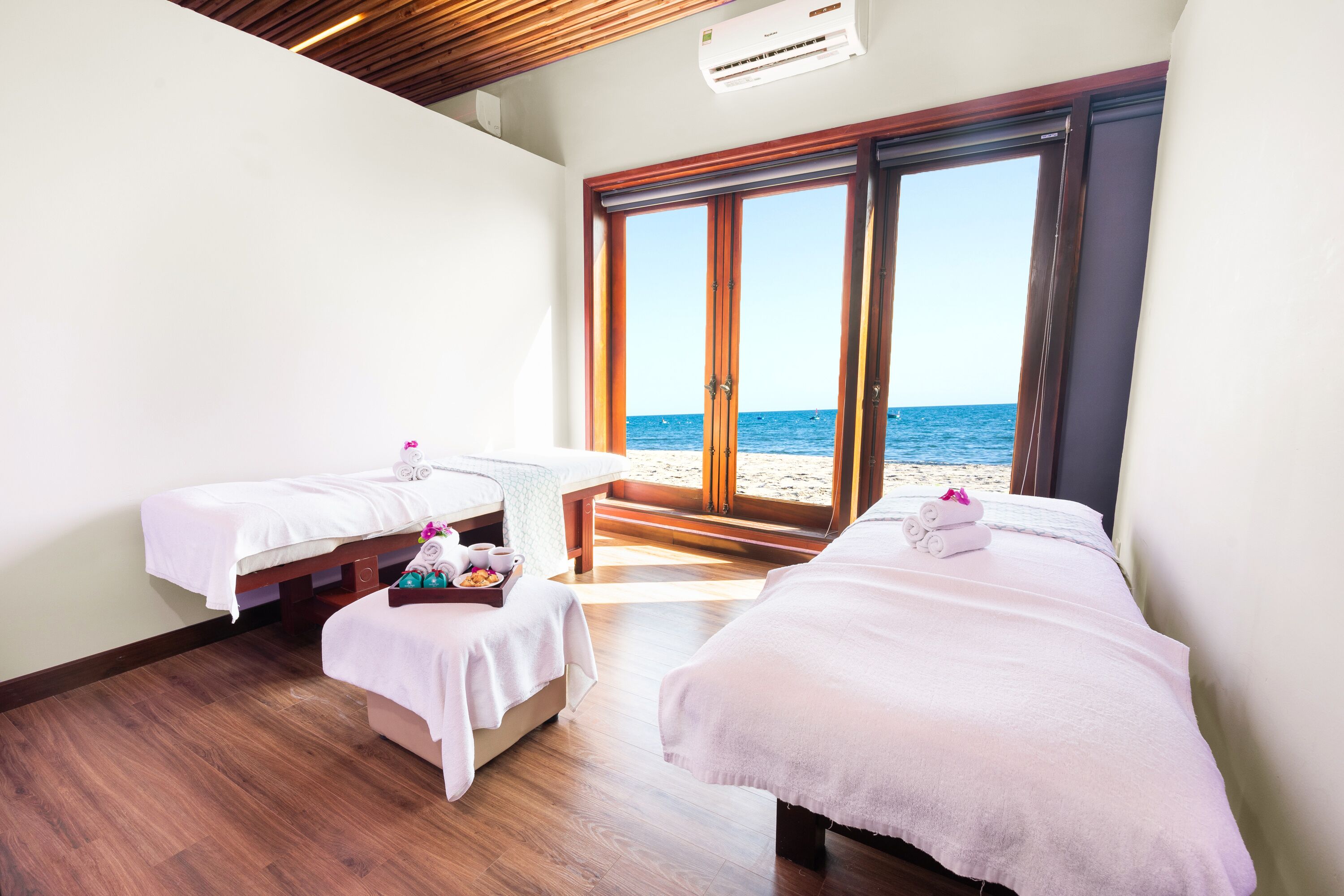 A room with two beds, a small table with a tray of snacks, and a view of the beach through large sliding doors.