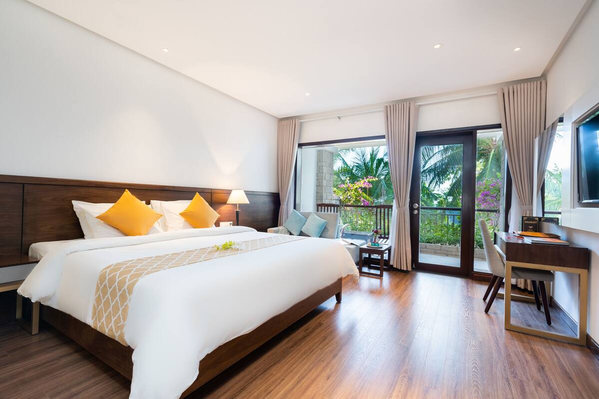 A hotel room with a large bed, a desk, and a balcony with a view of greenery.