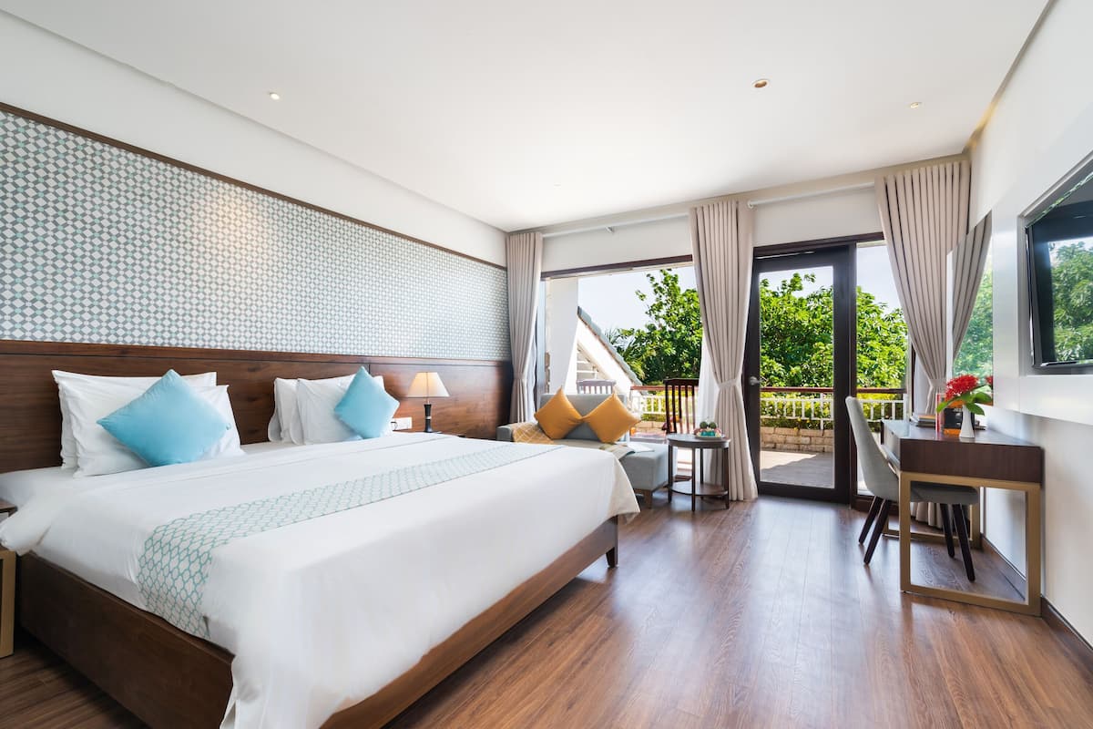 A modern hotel room with a large bed, a desk, and a balcony with a view of greenery.
