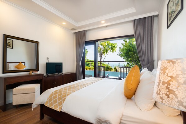 Habitación de hotel con cama, escritorio, silla, televisor y balcón con vistas a zonas verdes y un edificio.