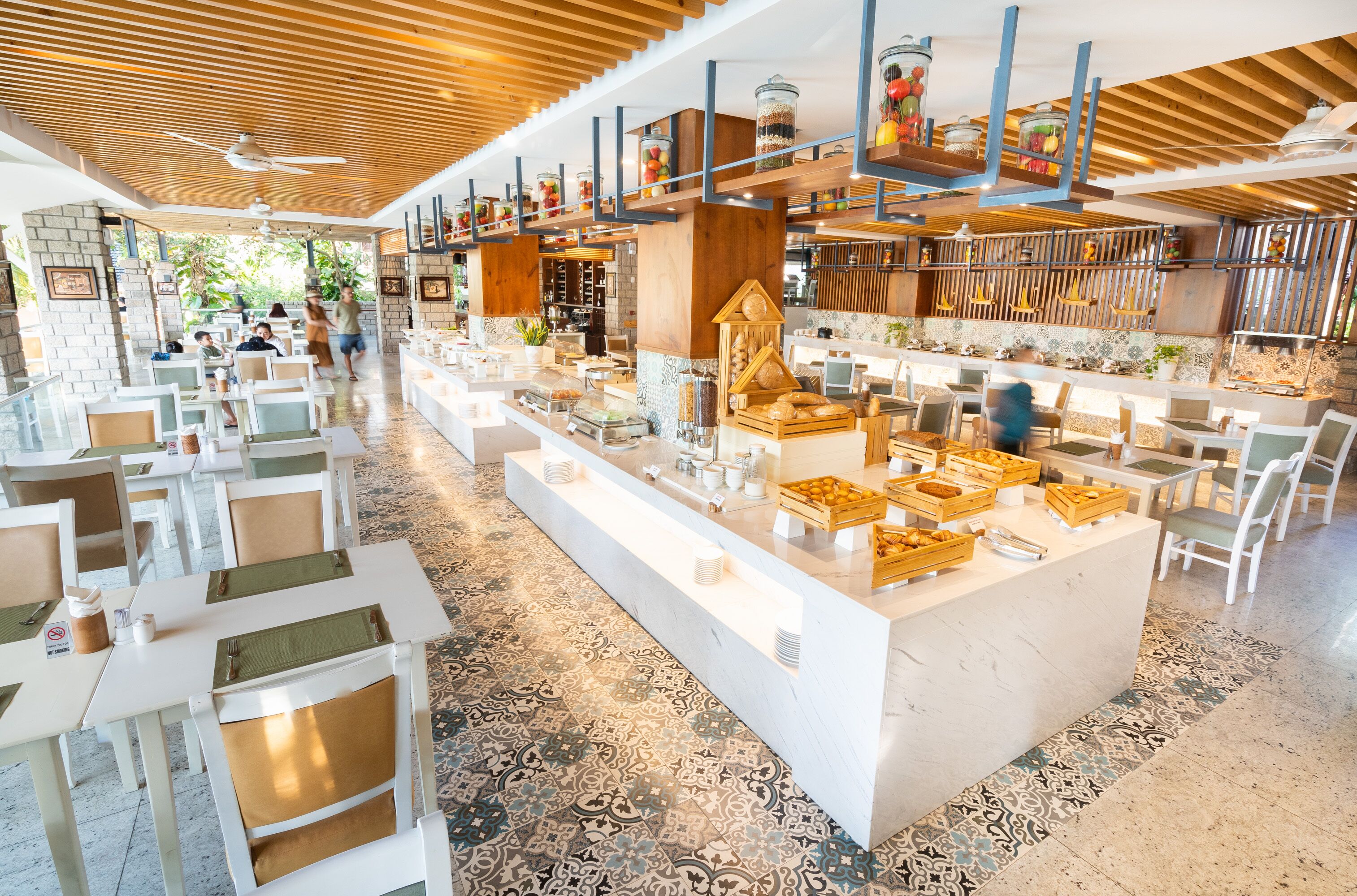 Um restaurante moderno com mesas e cadeiras brancas, um balcão central com expositores de comida e um teto com vigas de madeira.