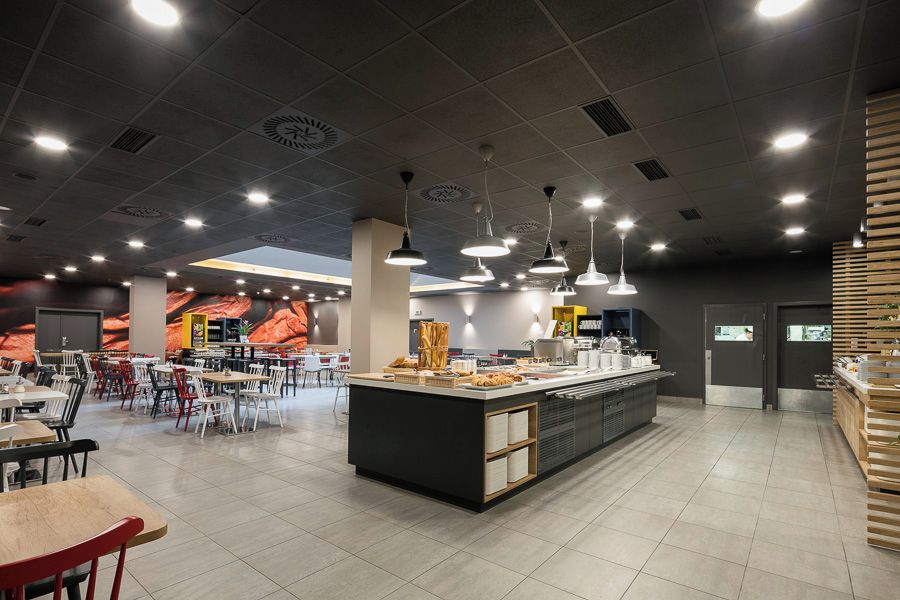 Une cafétéria moderne dotée de nombreuses tables et chaises, d’un comptoir de service central et d’un éclairage suspendu.