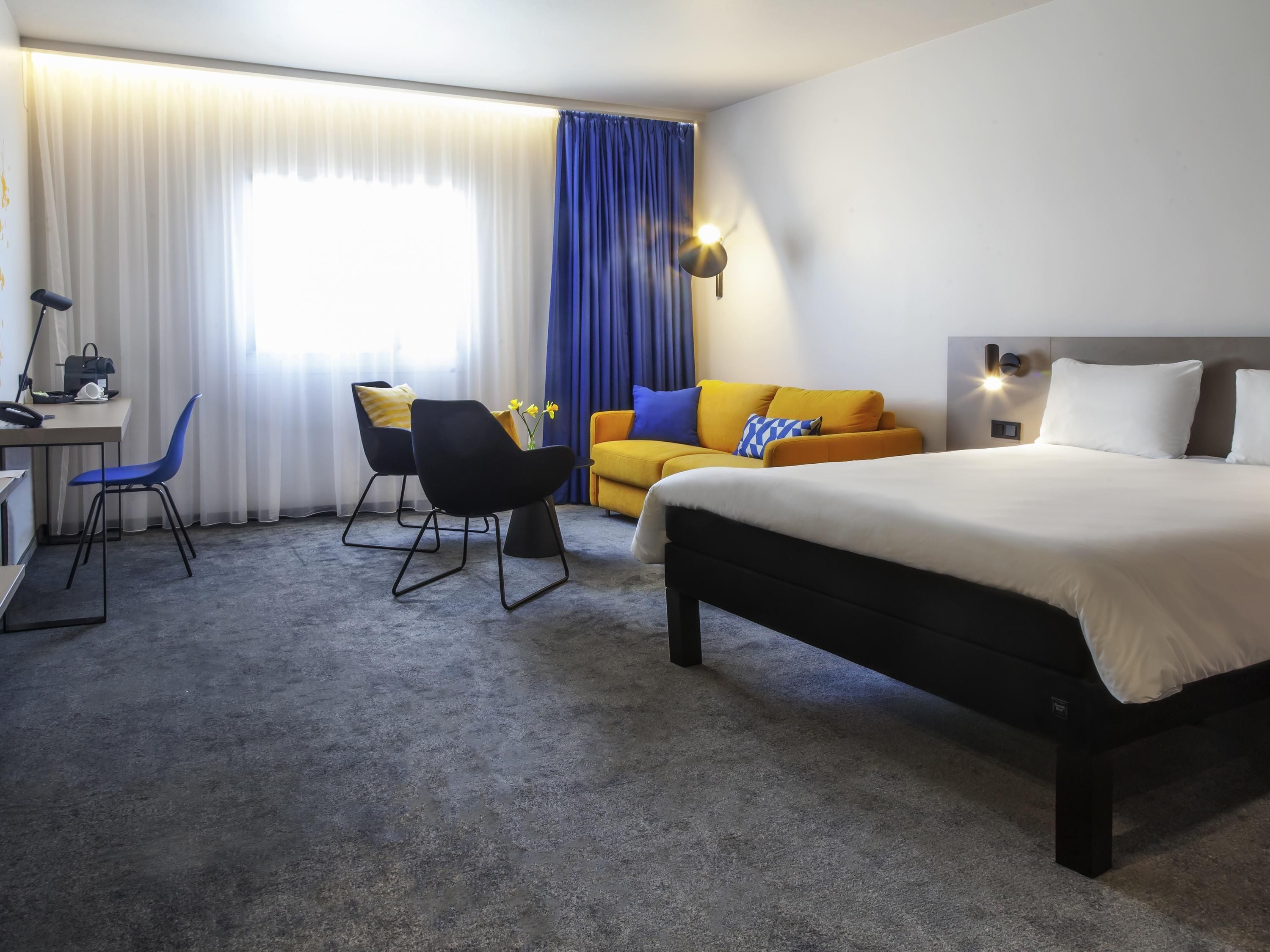 Une chambre d’hôtel avec un lit, un bureau, une chaise, un canapé et une fenêtre avec des rideaux.