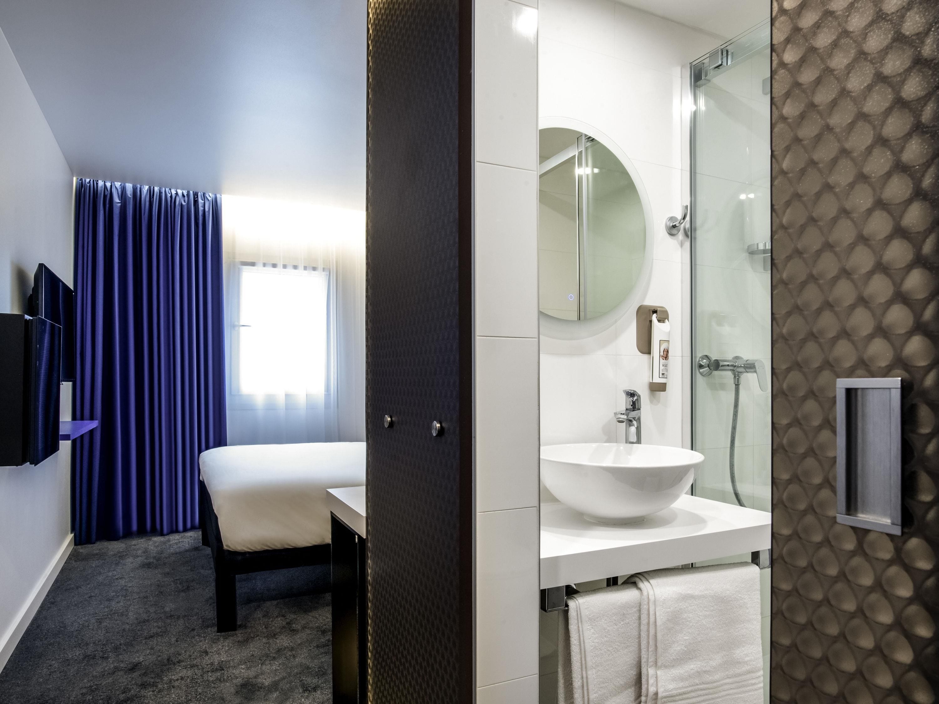A hotel room with a bed, a bedside table, a TV, a bathroom with a sink, a mirror, and a shower.