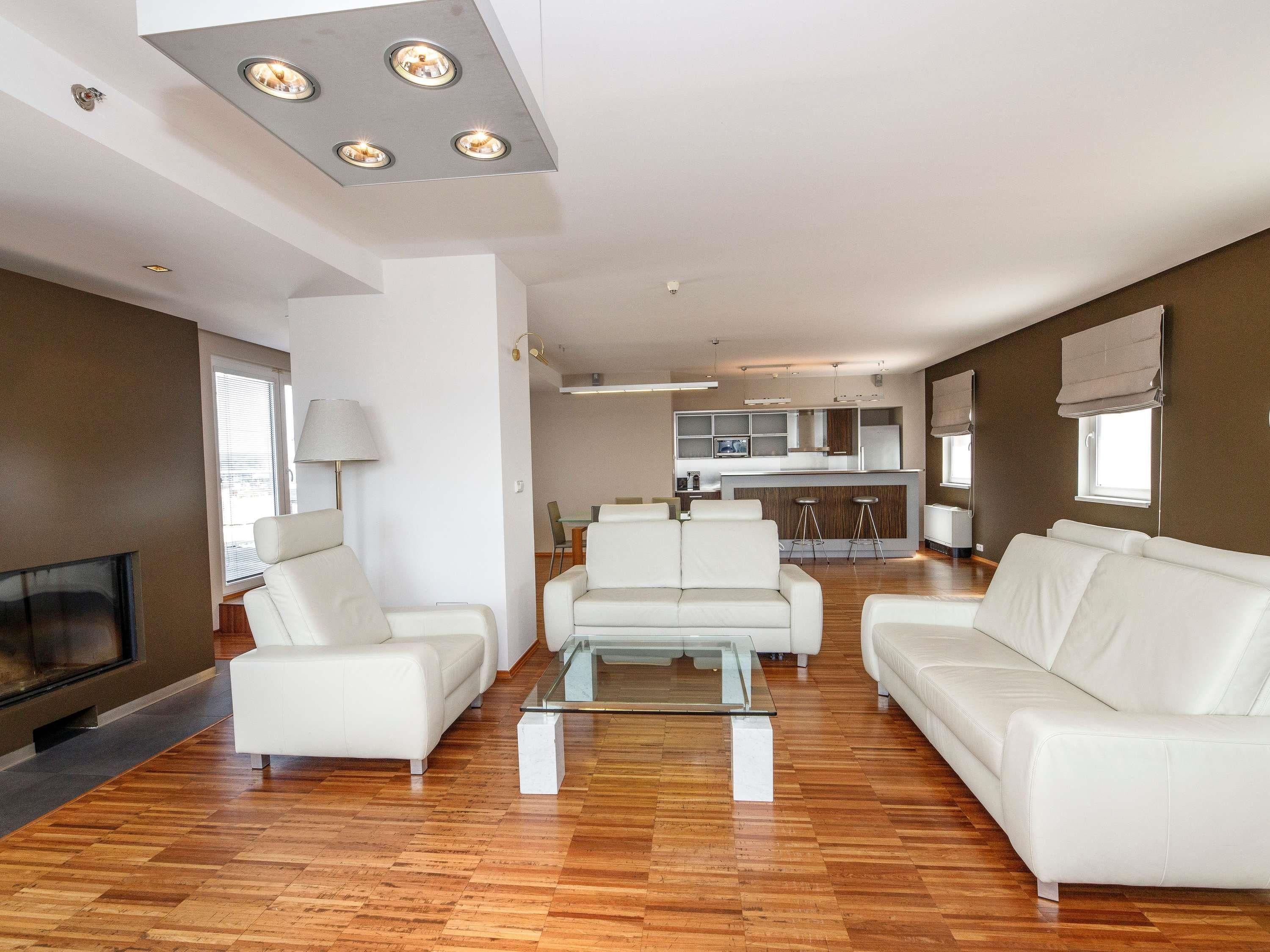 A modern living room with a TV, white sofas, a glass coffee table, and a kitchen in the background.