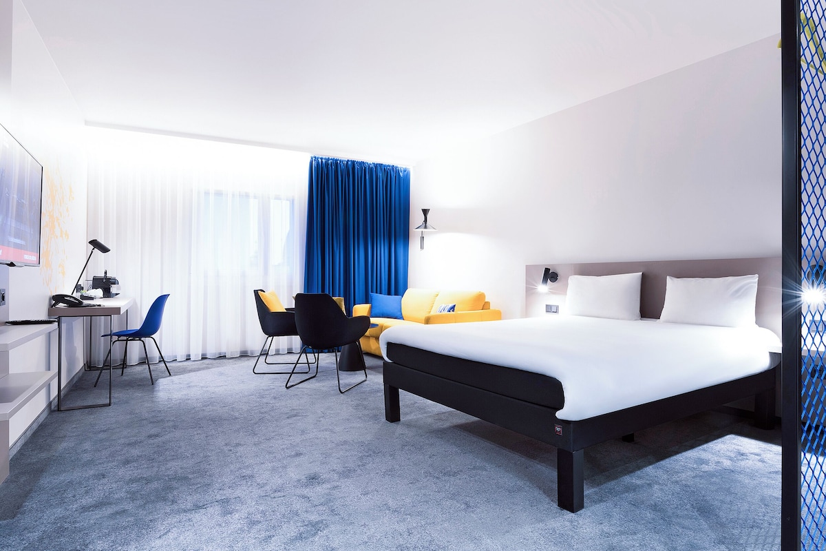 Una habitación de hotel con una cama, un escritorio, dos sillas, una lámpara y un televisor.