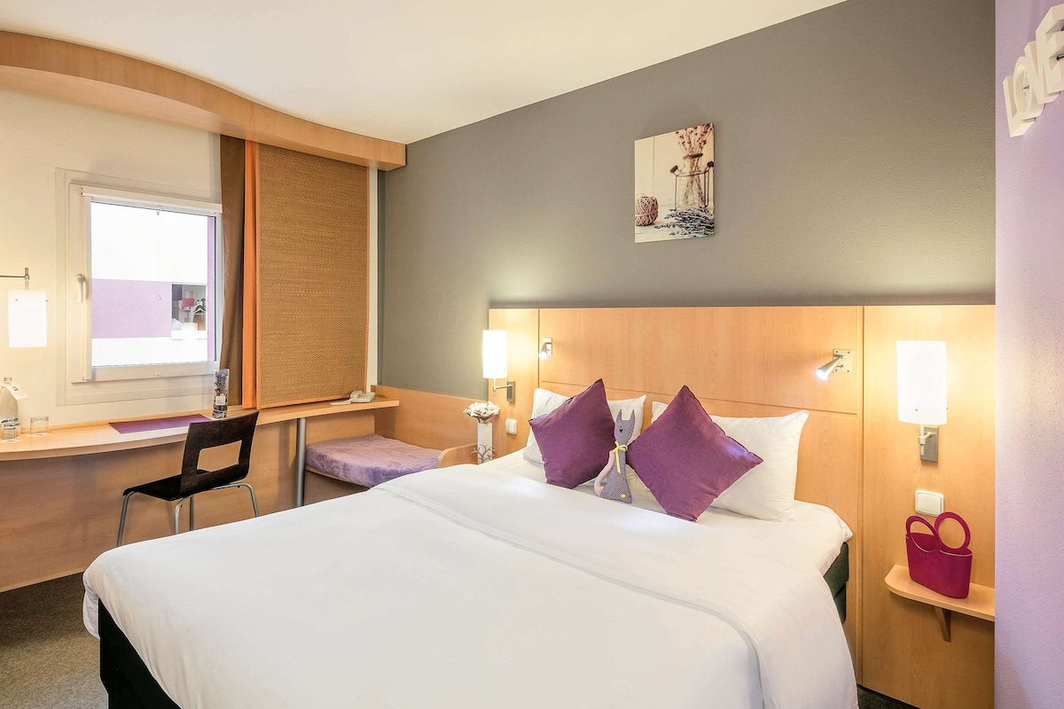 Una habitación de hotel con una cama, un escritorio, una silla y una mesita de noche con una lámpara.