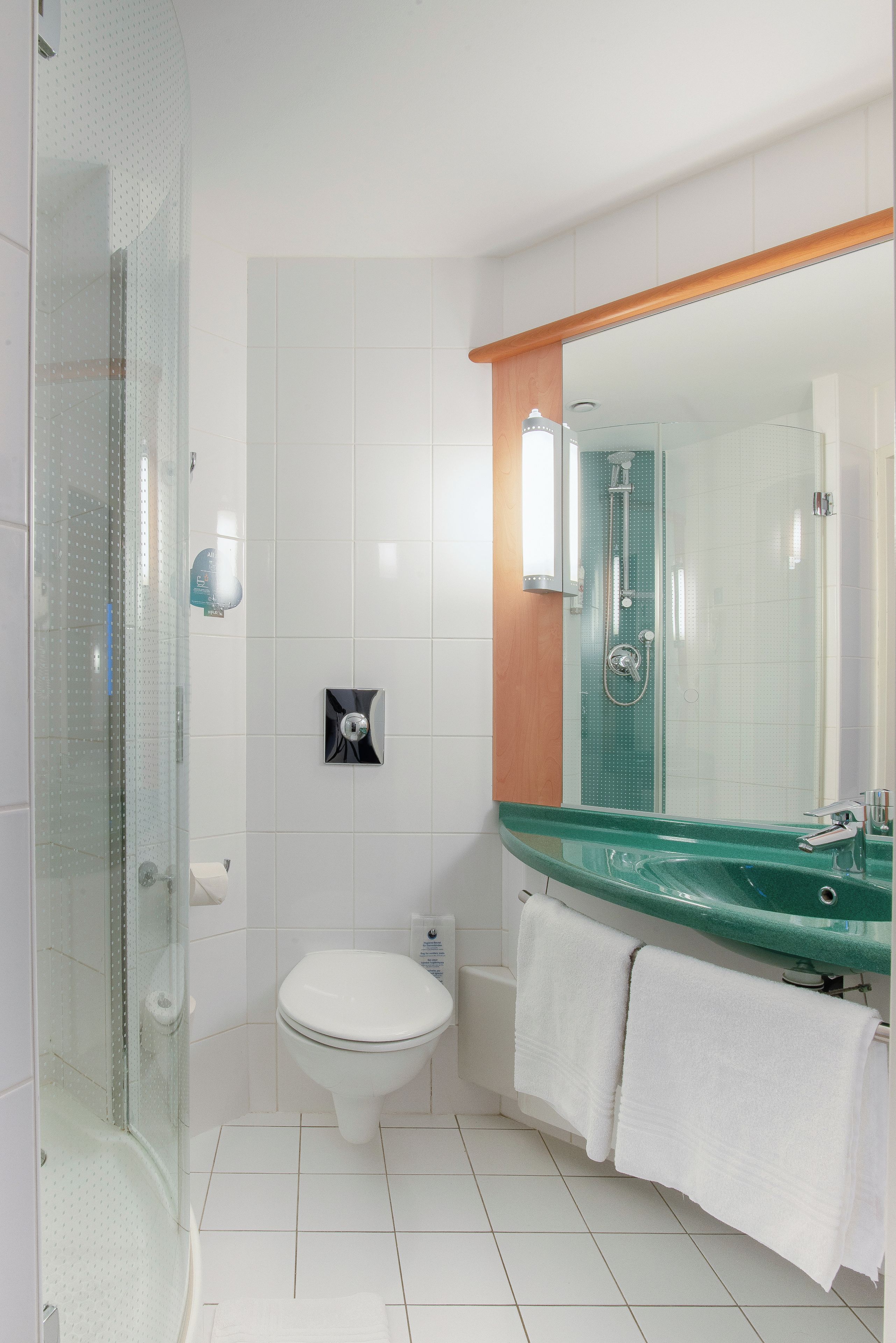 Un baño con cabina de ducha de vidrio, inodoro blanco, lavamanos verde con espejo y dos toallas blancas.