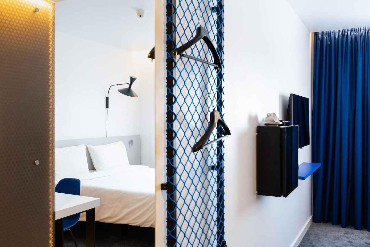 Habitación de hotel con cama, escritorio, silla, lámpara de escritorio, televisión y cortina.