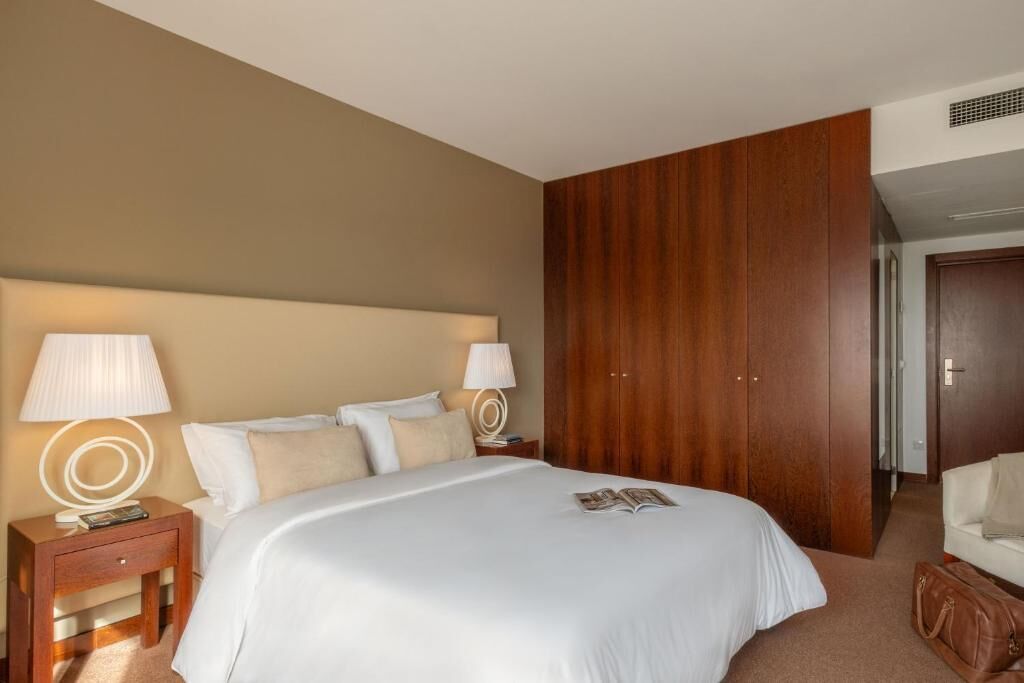 A hotel room with a large bed, two bedside tables with lamps, a wooden wardrobe, and a white armchair.