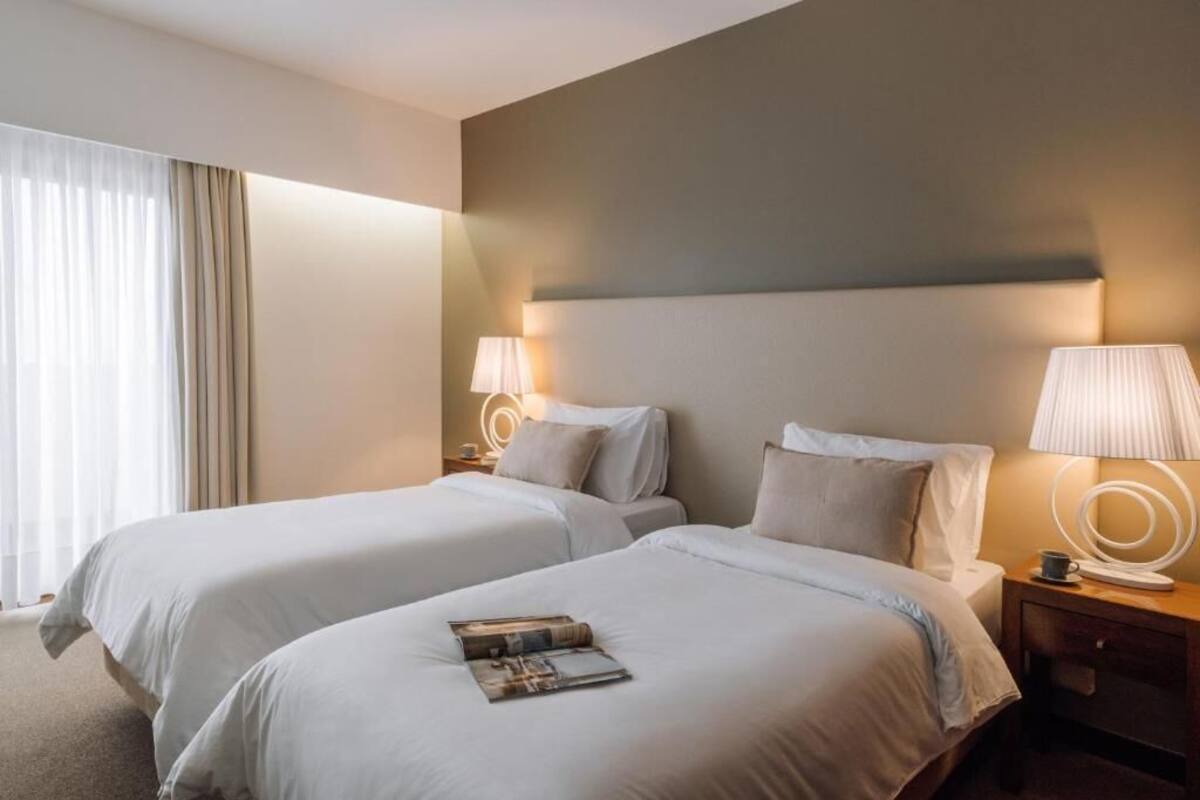 A hotel room with two beds, a nightstand with a lamp, and a window with curtains.