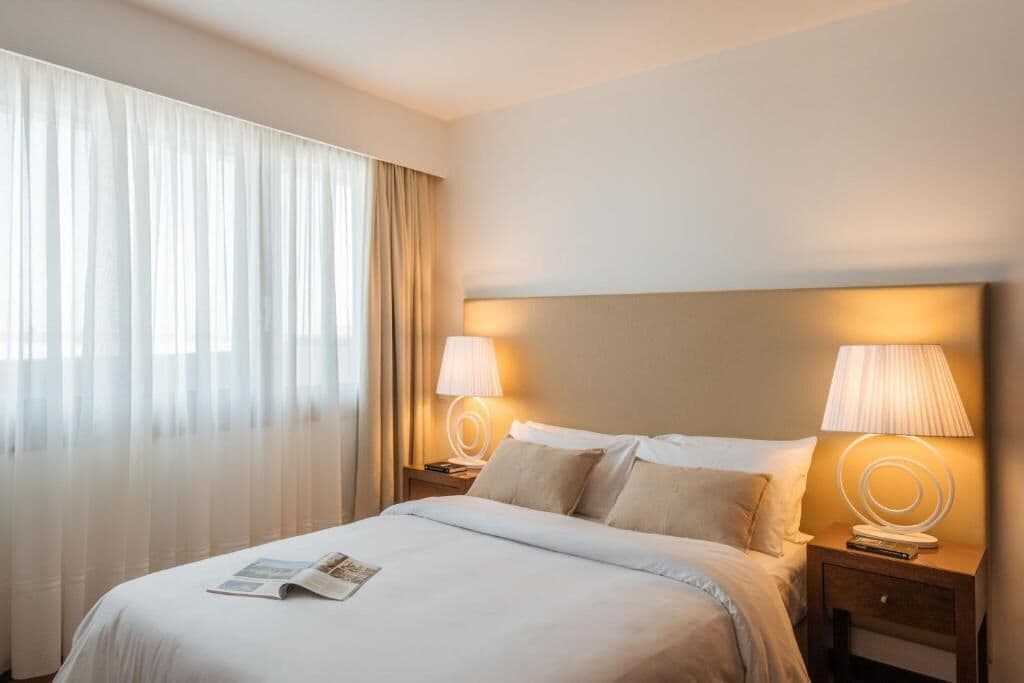 Habitación de hotel con una cama grande, dos lámparas de noche, una ventana con cortinas translúcidas y una revista sobre la cama.