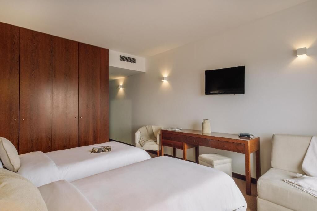 A hotel room with two beds, a wooden wardrobe, a desk with a chair, a television, and wall-mounted lights.