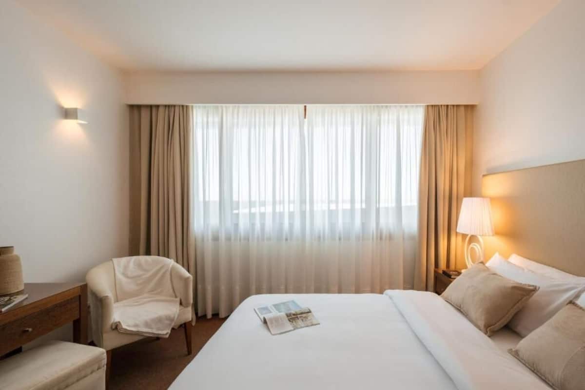 A hotel room with a large bed, a chair, a desk, and a nightstand with a lamp.
