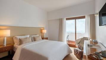 Quarto de hotel com uma cama grande, uma cadeira, uma escrivaninha com abajur e vista para o exterior.