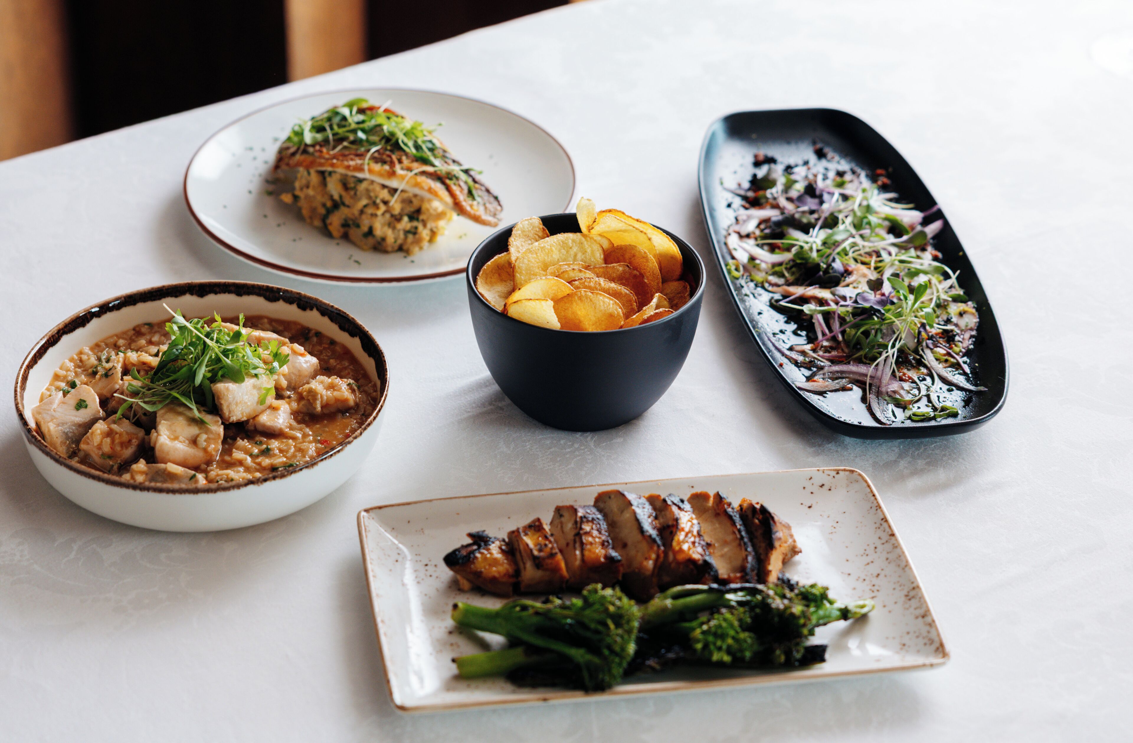 A table with various dishes including a bowl of chips, a plate of meat, and a dish with vegetables.