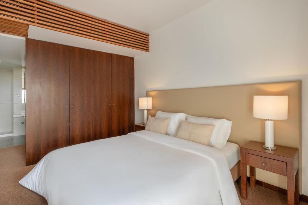A hotel room with a large bed, wooden wardrobe, two bedside tables with lamps, and a ceiling vent.