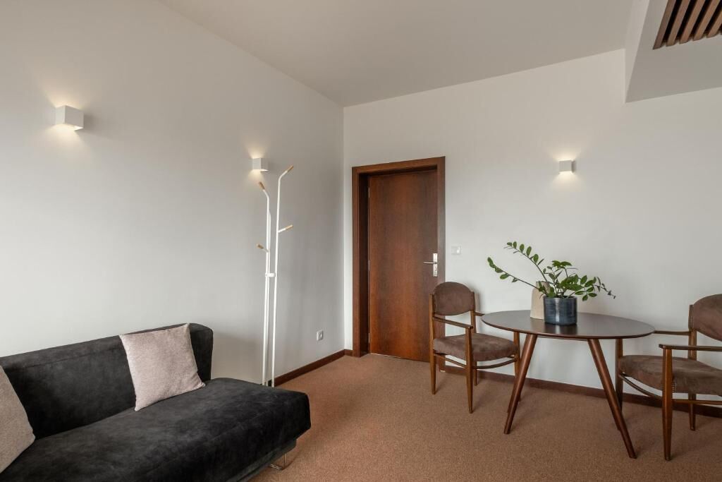 A modern living room with a dark sofa, a round wooden table, and two chairs. There is a wooden door and a floor lamp.