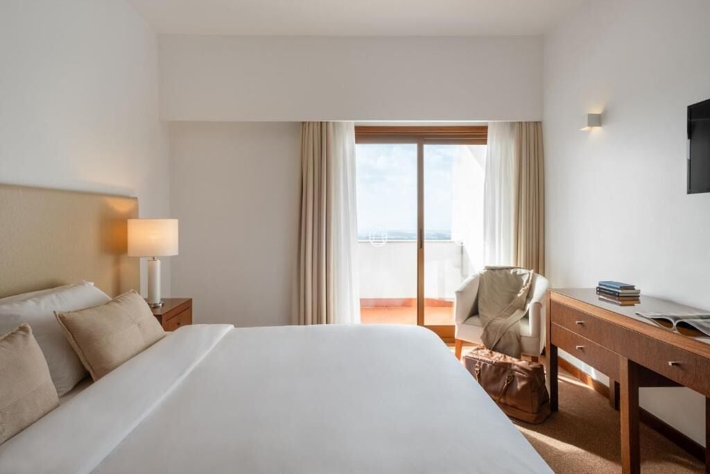 A hotel room with a bed, a desk, a chair, a lamp, and a view through a window.