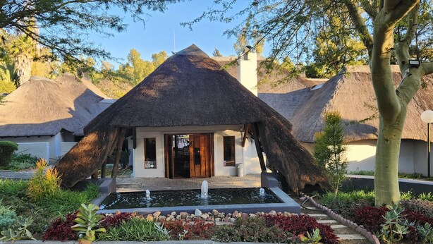 A thatched-roof building with a water feature in front, surrounded by landscaped gardens. - Shumba Valley Lodge Lanseria Airport (Lanseria)