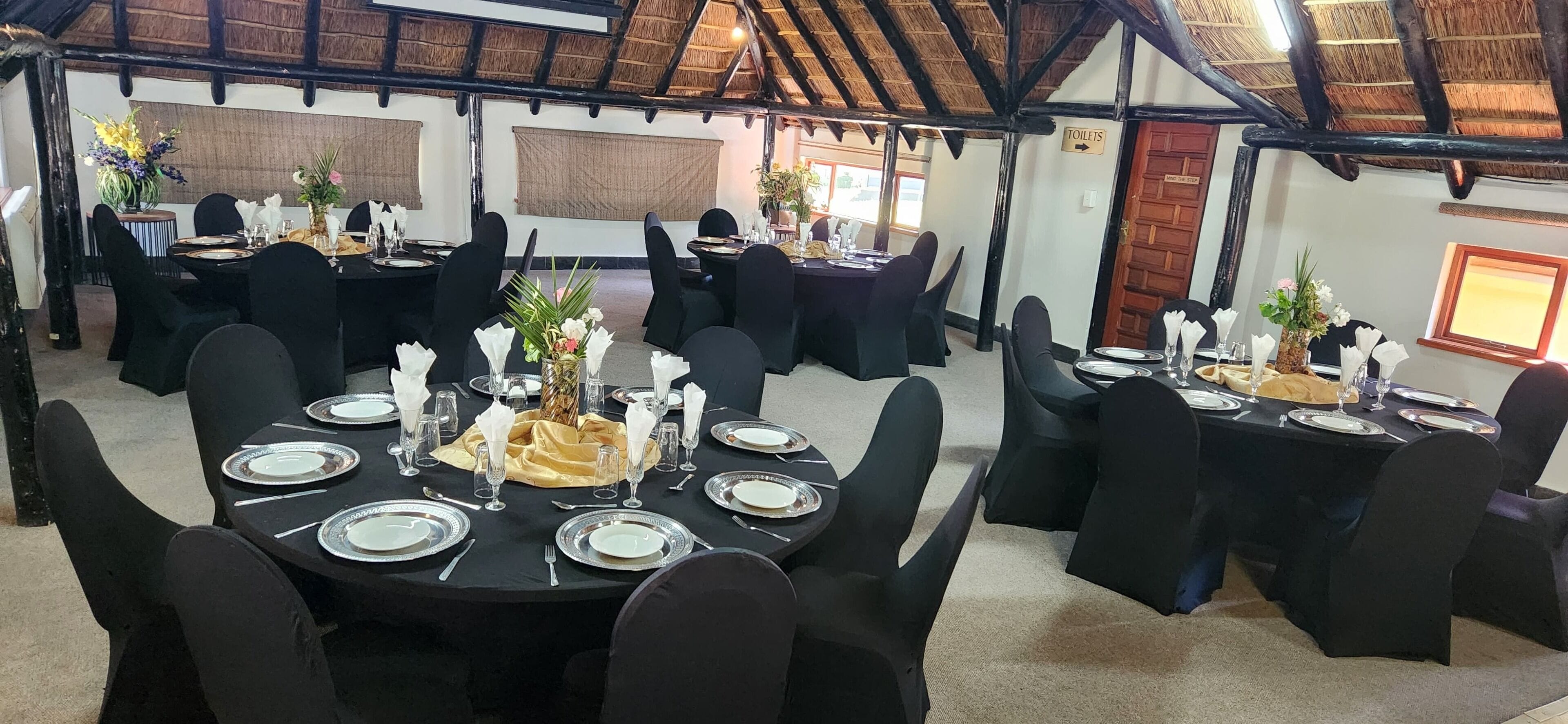 Uma área de refeições com mesas redondas preparadas para vários convidados, com cadeiras pretas e louças brancas.