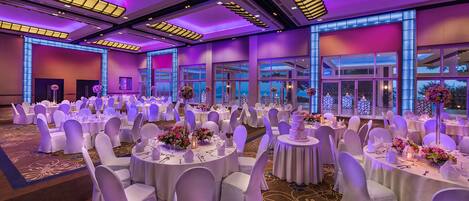 Un salón de banquetes con mesas redondas, sillas blancas e iluminación morada.