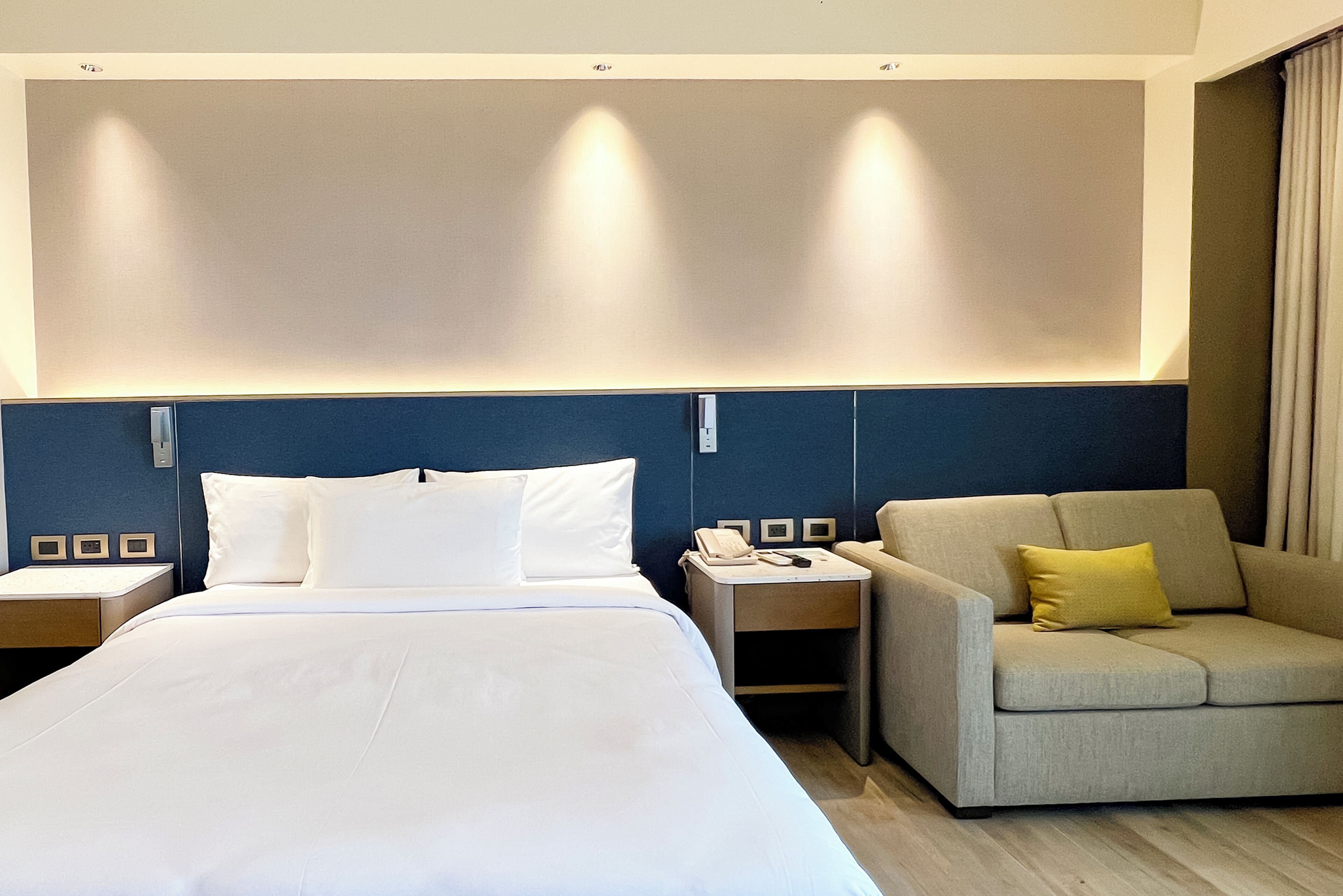 A hotel room with a bed, a sofa, a nightstand, and a wall-mounted phone.