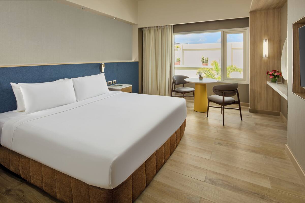 A hotel room with a large bed, a small table, two chairs, a window with curtains, and a view of the outside.