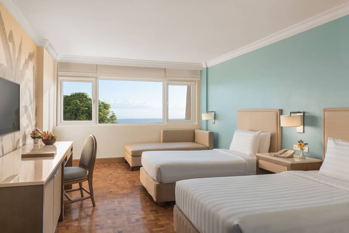 A hotel room with two beds, a desk, a chair, a TV, and a window with a view of the outdoors.