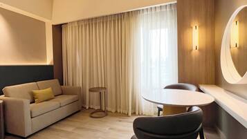 A hotel room with a sofa, a round table, a chair, a small stool, and a large window with curtains.