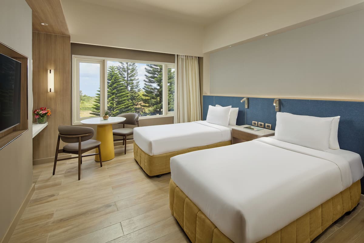 A hotel room with two beds, a dining table, a TV, and a window with a view of trees.