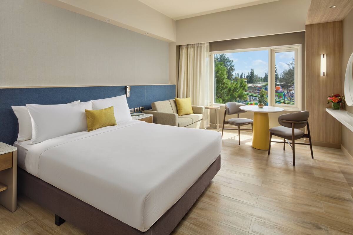 A hotel room with a bed, a small table, two chairs, a nightstand, and a window with a view of a playground.