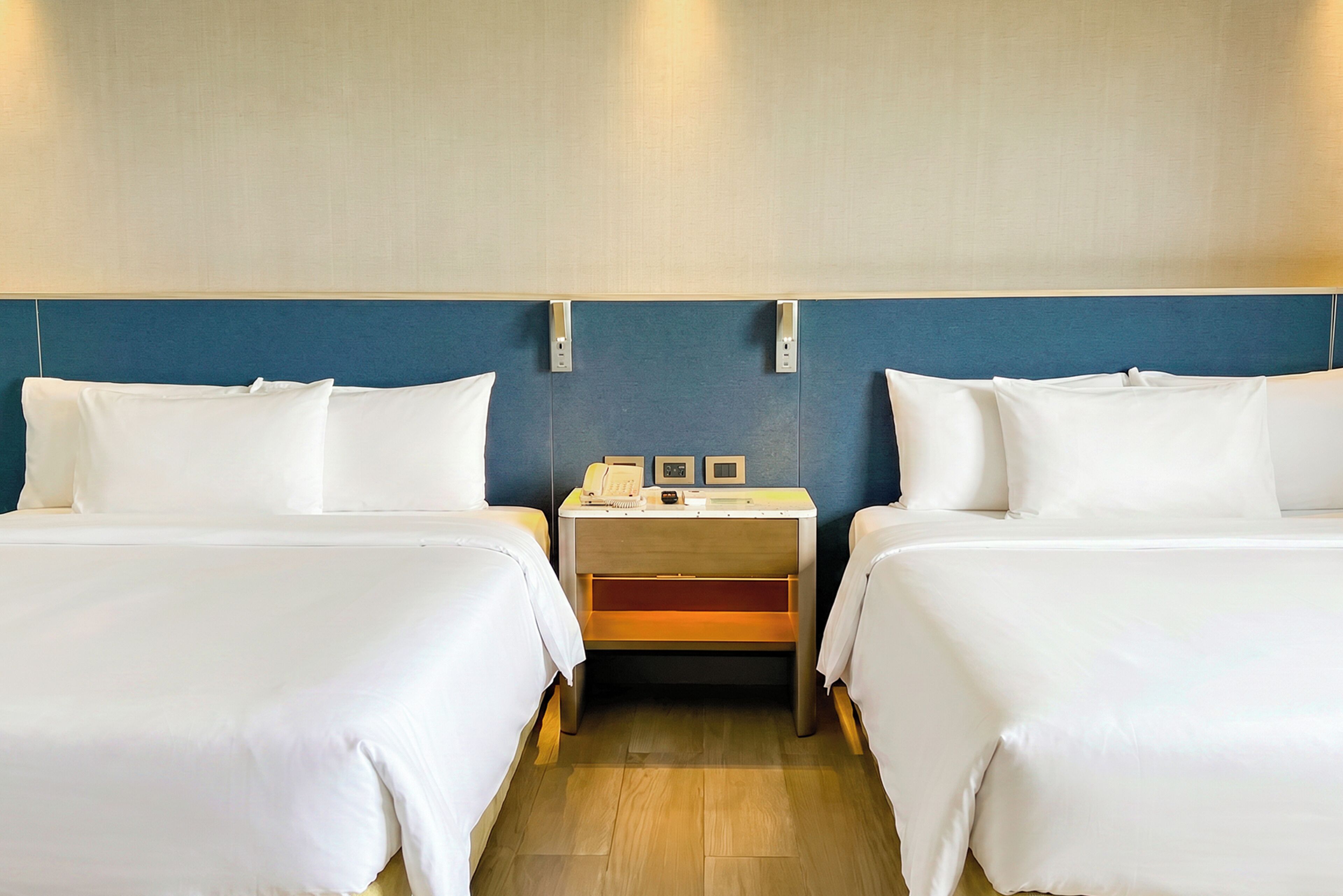 Une chambre d’hôtel avec deux lits simples, une table de chevet avec une lampe, une petite table avec un téléphone et une horloge, et un luminaire fixé au mur.