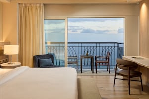A hotel room with a bed, a desk, two chairs, a small table, and a balcony with a view of the sea.