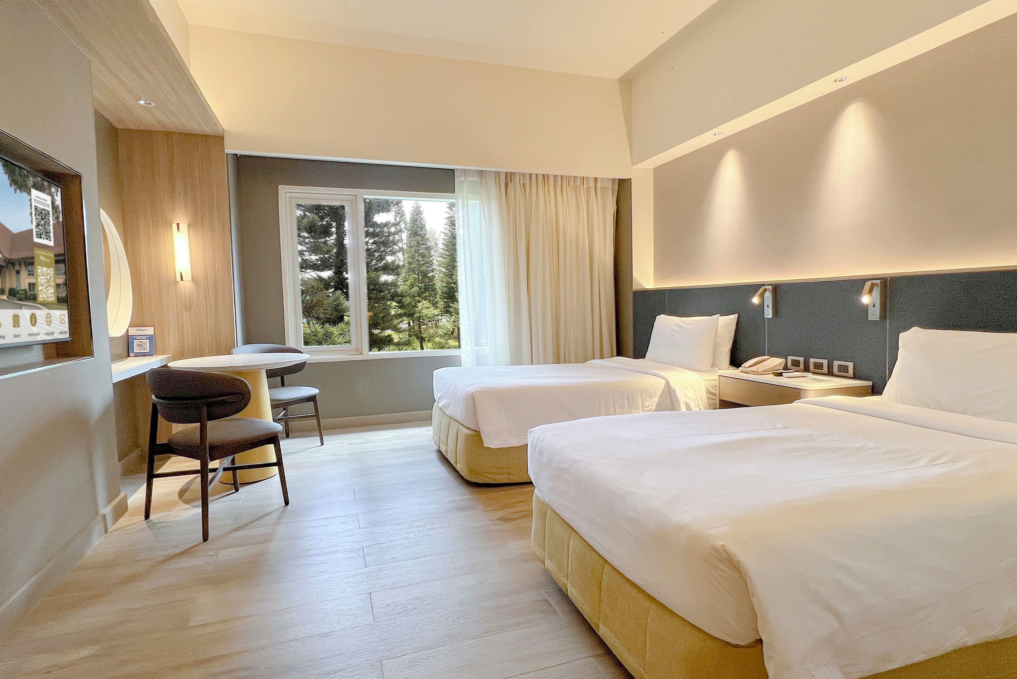 Ein Hotelzimmer mit zwei Betten, einem Schreibtisch und einem Fenster mit Blick auf Bäume.