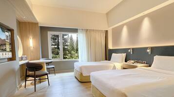 A hotel room with two beds, a desk, and a window with a view of trees.