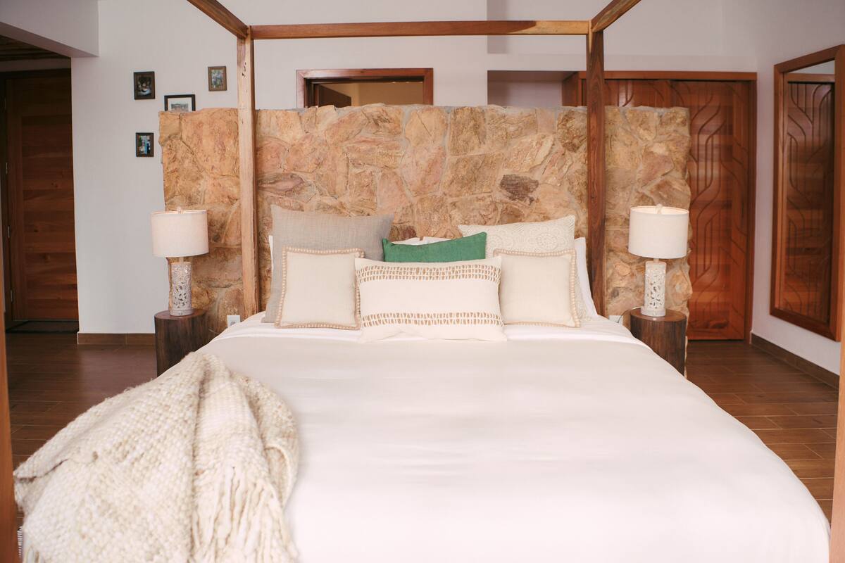 Un dormitorio con cabecera de piedra, una cama grande con muchos cojines, dos lámparas de noche y suelo de madera.