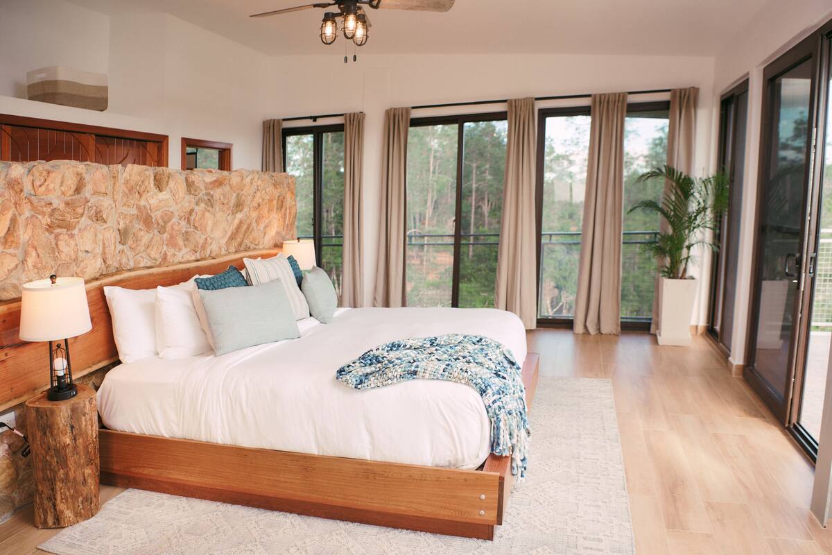 Un dormitorio amplio con una cama grande, cabecera de madera y vistas al exterior a través de amplios ventanales.