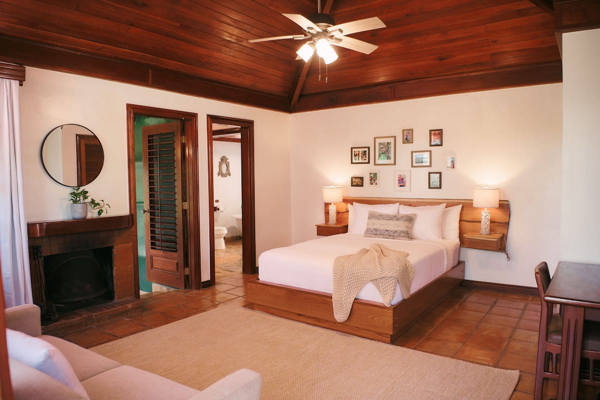 Un dormitorio con techo de madera, una cama con cubrecama blanco, dos mesitas de noche con lámparas, una chimenea y un espejo en la pared.