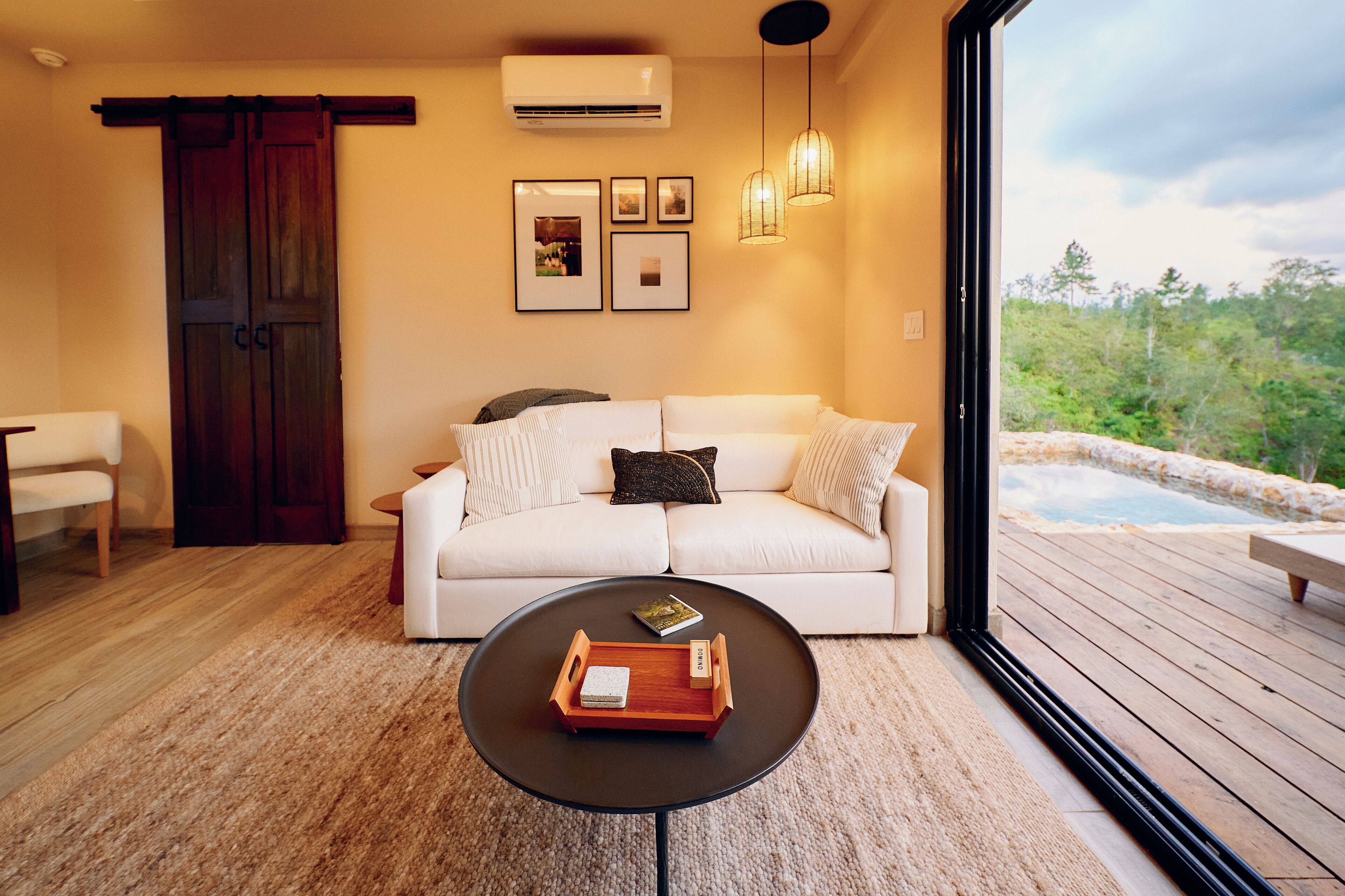 A modern living room with a white sofa, a round coffee table with a tray, and a large window overlooking a natural landscape.