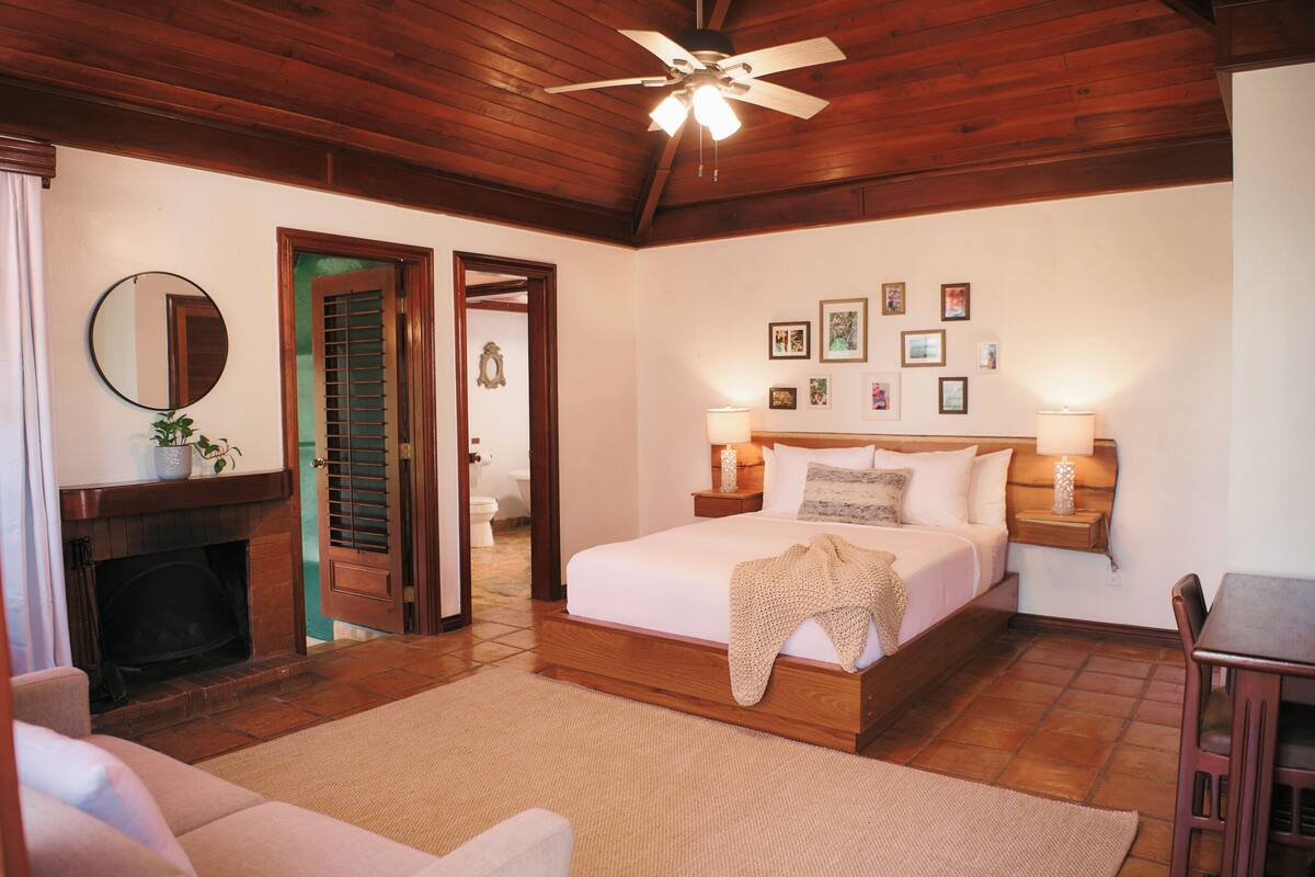 Un dormitorio con techo de madera, una cama con cubrecama blanco, dos mesitas de noche con lámparas, una chimenea y un espejo en la pared.
