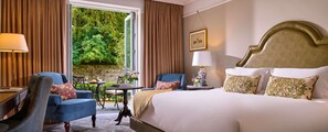 A hotel room with a large bed, two armchairs, a small table, and a view of greenery through a window.
