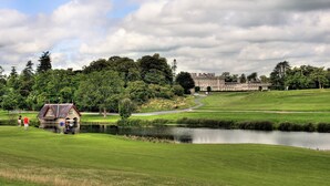 A golf course with a pond, a small house, and a large building in the background.