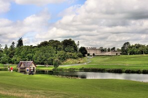Um campo de golfe com um lago, uma pequena casa e um edifício grande ao fundo.
